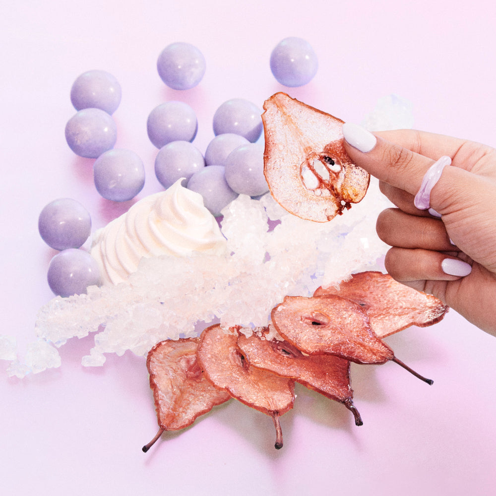 Hand holding a slice of pear over a dessert with purple grapes and whipped cream on a pink background