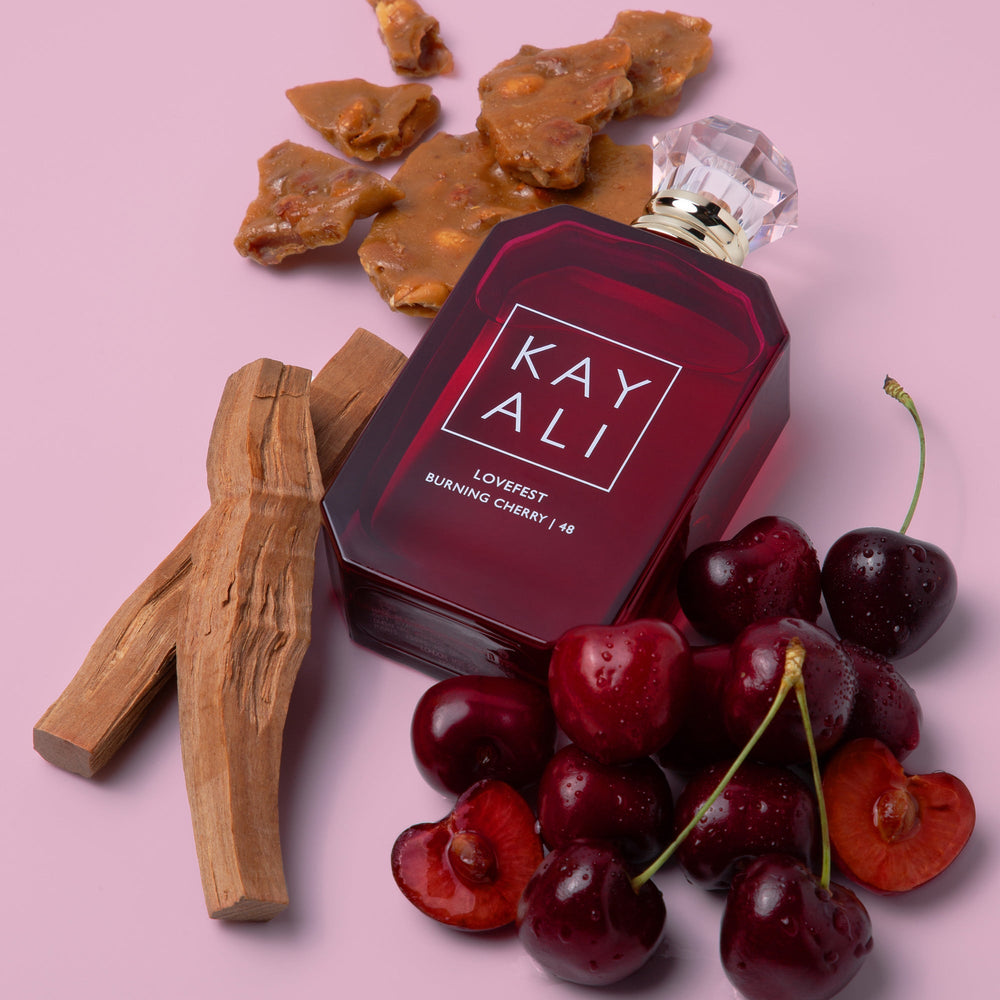 Red perfume bottle labeled 'KAY ALI' with cherries and wooden elements on a pink background