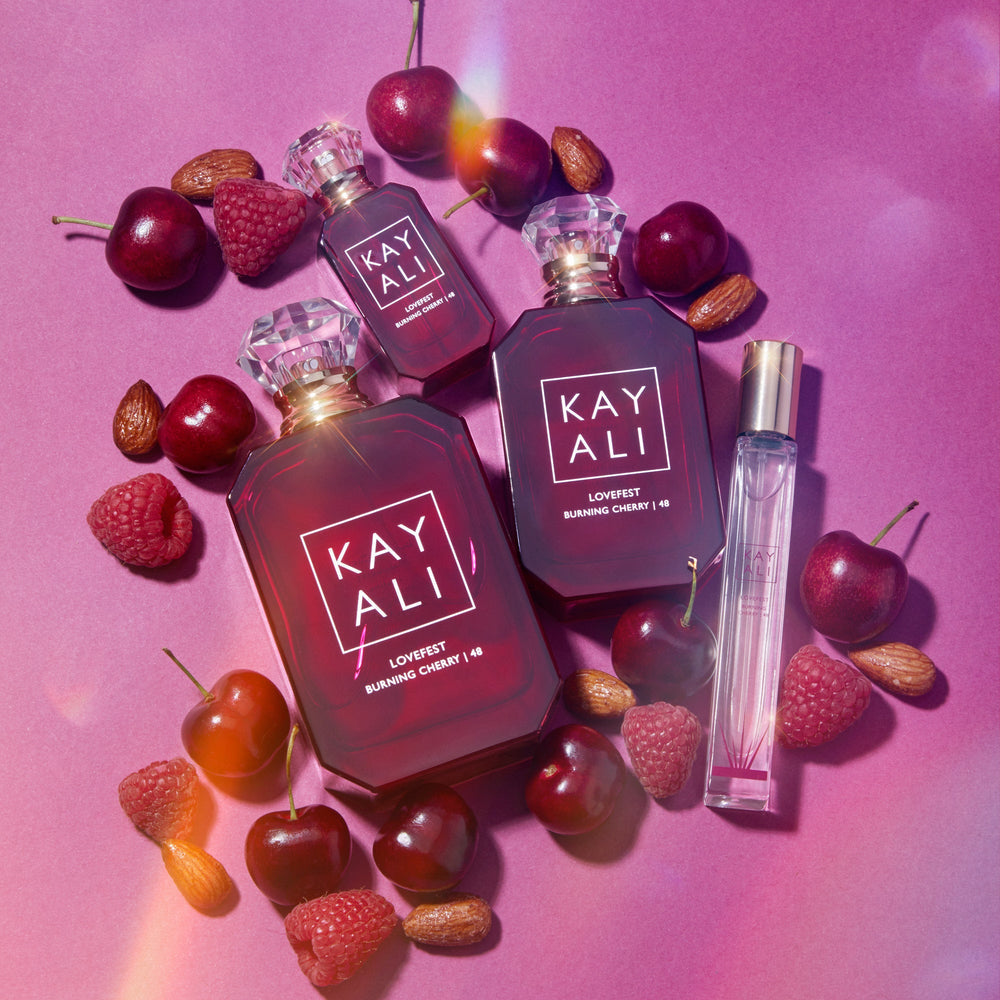 Three bottles of Kayali perfume surrounded by cherries and raspberries on a pink background