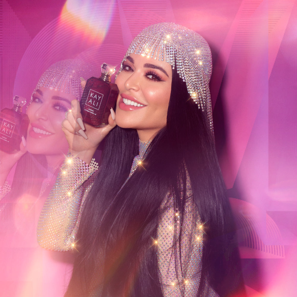 Woman holding a Kay Ali perfume bottle with a sparkling headpiece against a pink background