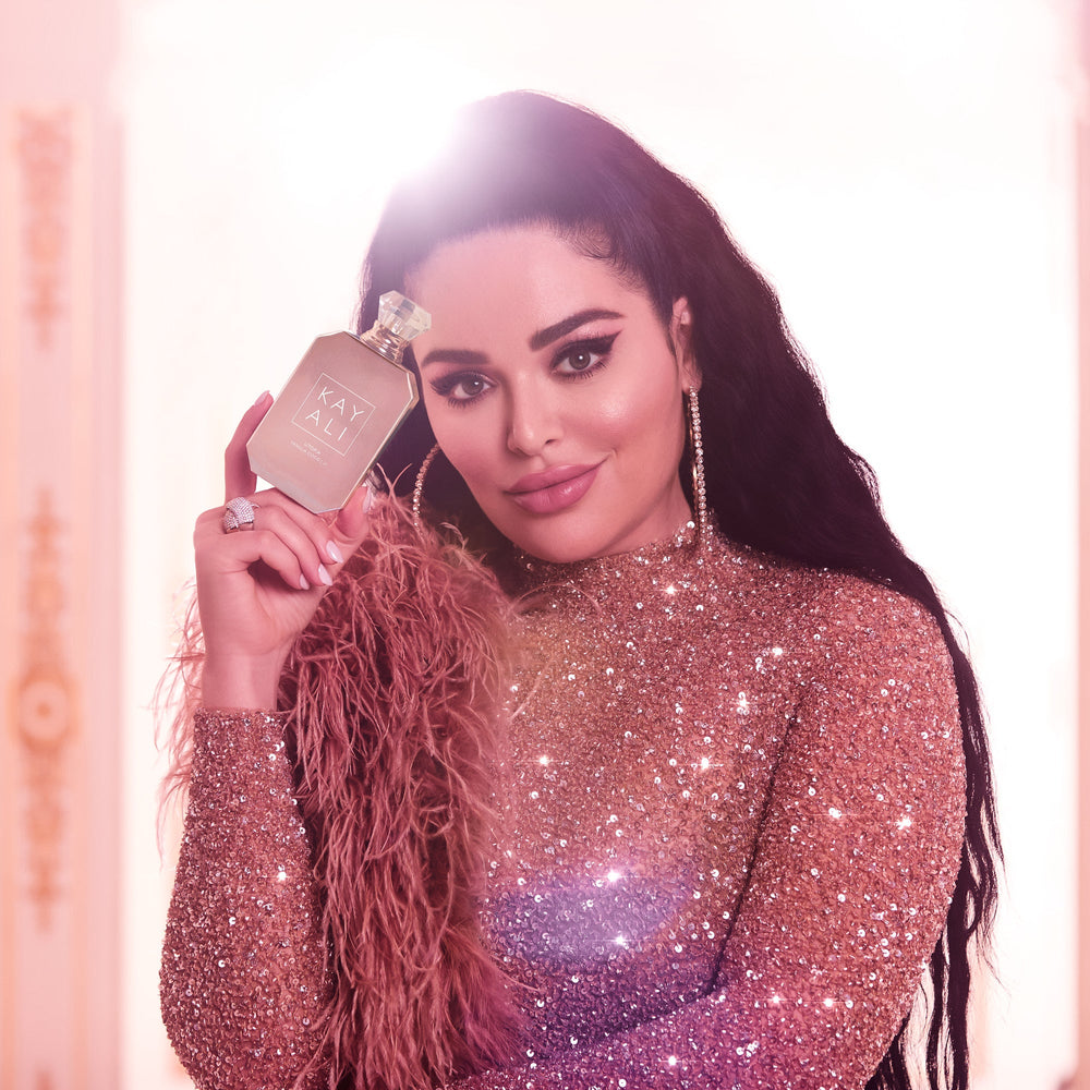 Woman in a glittery outfit holding a perfume bottle with a pinkish-beige background